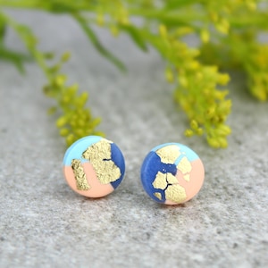 May include: A pair of small, round stud earrings with a blue, pink, and gold design. The earrings are made of polymer clay and have a matte finish.