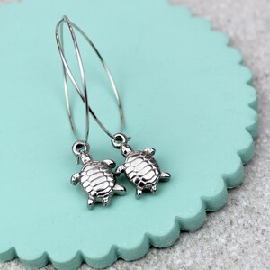 May include: Silver-coloured hoop earrings featuring small turtle charms. The earrings are presented on a light blue, scalloped-edge surface against a grey background. The turtle charms have detailed shell patterns.