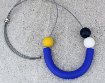 Può includere: Una collana con un lungo ciondolo a tubo blu curvo, una perla sferica gialla, una perla sferica bianca e una perla sferica blu scuro. Il ciondolo è sospeso su un sottile cordino grigio con una perlina di regolazione in metallo color argento.