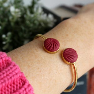 May include: Gold-coloured cuff bracelet with two circular, burgundy-coloured accents with a textured, pleated design. The bracelet is open, allowing it to be slipped onto the wrist. The background is blurred, with a hint of greenery.