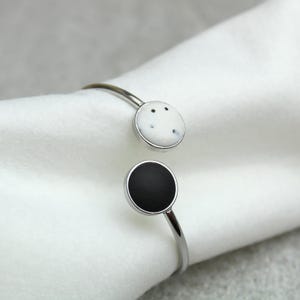 Könnte beinhalten: Ein silberfarbenes offenes Armreif-Armband. Das Armband hat zwei kreisförmige Akzente: einen weißen mit schwarzen Sprenkeln und einen anderen in schlichtem Schwarz. Das Armband wird auf einem weißen Stoff präsentiert.