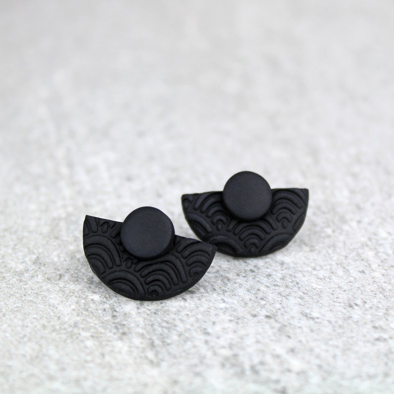 May include: A pair of black stud earrings with a half-circle design and a textured pattern. The earrings are made of polymer clay.