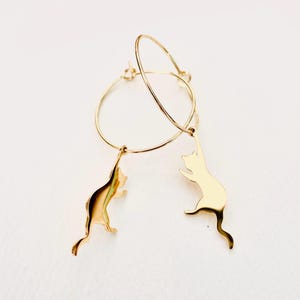 May include: Gold hoop earrings with dangling cat silhouettes. The cats are in a jumping pose.