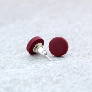 May include: A pair of small, round, burgundy stud earrings. The earrings are made of a smooth, matte material and have a silver post.