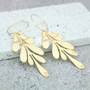 May include: Gold hoop earrings with a dangling leaf design. The leaves are cut out and have a textured finish.