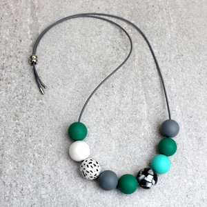 May include: A contemporary necklace with a grey cord and a selection of colourful, spherical beads. The beads include shades of green, white, grey, and a patterned black and white bead. The necklace is displayed on a grey surface.