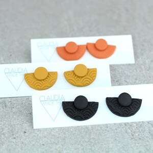 May include: Three pairs of earrings with a half-circle design and a textured pattern. The earrings are in black, orange, and yellow.