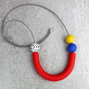 May include: A necklace featuring a long, curved red tube, a white bead with black spots, a solid blue sphere, and a solid yellow sphere strung on a grey cord. The cord includes two small silver-toned metal beads for adjustment and a silver-toned clasp.