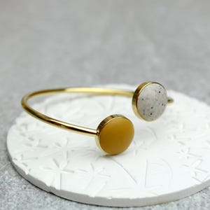 Könnte beinhalten: Ein goldfarbenes, offenes Armreif-Armband mit zwei kreisförmigen Akzenten. Einer ist gesprenkelt grau, der andere einfarbig senfgelb. Das Armband wird auf einer weißen, strukturierten Oberfläche präsentiert.