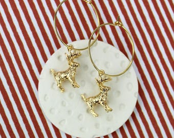 May include: Gold-colored hoop earrings with small, detailed deer charms. The earrings are displayed on a white, textured surface against a background of red and white diagonal stripes. The deer charms add a festive touch.
