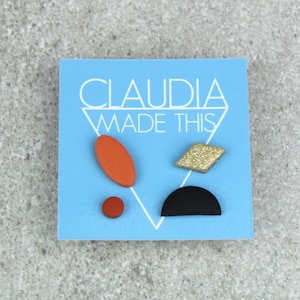 May include: A set of four colourful geometric earrings on a blue card with the text "CLAUDIA MADE THIS". The earrings are a red oval, a red circle, a gold glitter diamond, and a black half-circle.