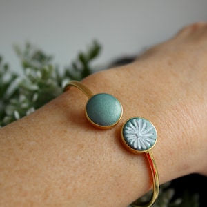 Könnte beinhalten: Goldfarbenes Armreif-Armband mit zwei dekorativen Elementen. Ein Element ist ein einfarbiger, teal-farbener Kreis, das andere ein teal-farbener Kreis mit weißem Blumenmuster. Das offene Armband liegt auf einem Handgelenk.