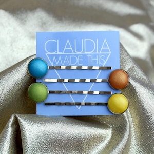 May include: Four bobby pins with colourful, round, metallic accents in blue, green, orange, and yellow. The pins are displayed on a light blue card that reads "CLAUDIA MADE THIS".