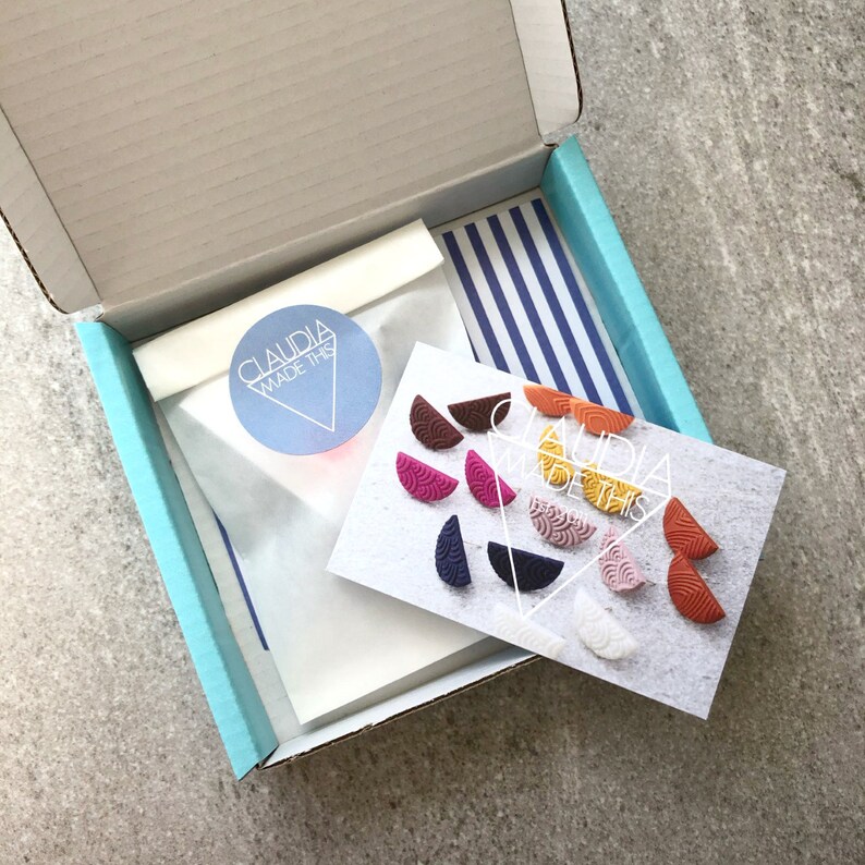 May include: A blue box with a white tissue paper bag inside. The bag has a round sticker that says "Claudia Made This". The box also contains a card with a variety of colorful half-circle earrings and the text "Claudia Made This".
