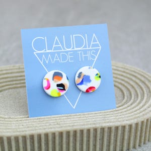 May include: A pair of white round stud earrings with a colorful abstract design. The earrings are displayed on a blue card with the text "CLAUDIA MADE THIS".