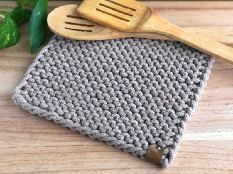 May include: A gray knitted pot holder with a woven texture. The pot holder is made of cotton rope and has a small brown leather tag.