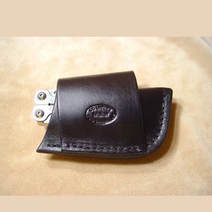 May include: A dark brown leather sheath with visible stitching along the bottom edge. The leather is embossed with an oval logo containing the text CC Leather Maker. A silver metal multi-tool is partially visible inside the leather pouch.