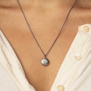 Tiny antiqued silver locket hangs on a 20 inches long antique silver chain. The model is wearing a cream color button down jersey tee.