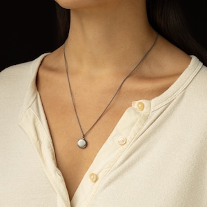 Tiny antiqued silver locket hangs on a 20 inches long antique silver chain. The model is wearing a cream color button down jersey tee.