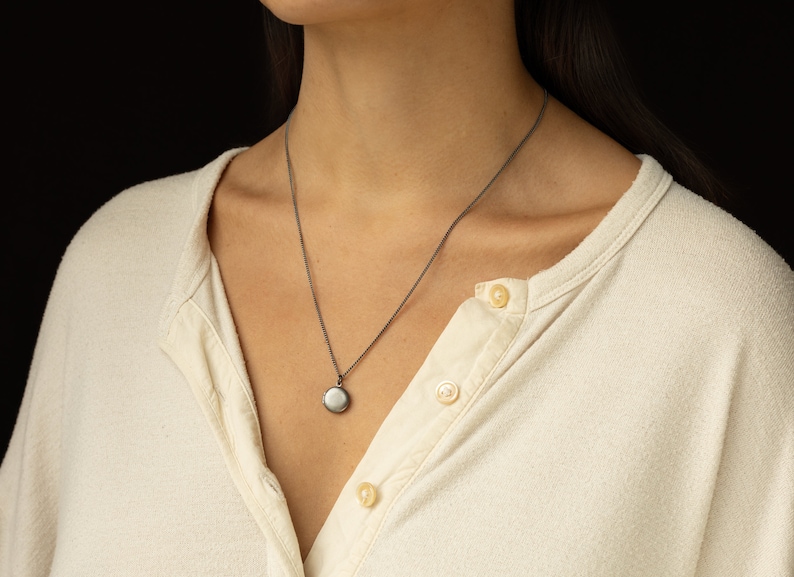 Tiny antiqued silver locket hangs on a 20 inches long antique silver chain. The model is wearing a cream color button down jersey tee.