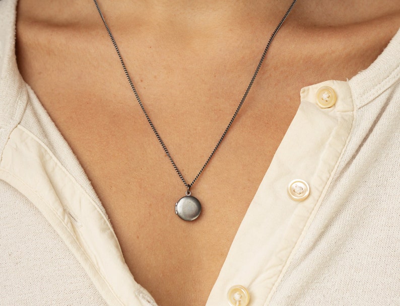 Tiny antiqued silver locket hangs on a 20 inches long antique silver chain. The model is wearing a cream color button down jersey tee.