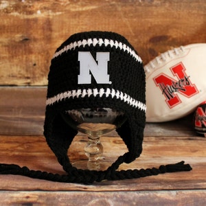 May include: Black and white crocheted hat with ear flaps. The hat features a white "N" logo on the front and white stripes. A white and red football with the word "Huskers" is in the background.