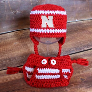May include: A red and white crocheted baby hat and diaper cover set. The hat has a white "N" and white stripes. The diaper cover has white stripes and two white buttons. The set is displayed on a wooden surface.