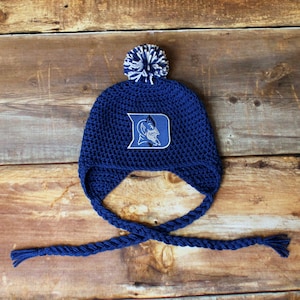 May include: A navy blue crocheted hat with ear flaps and a braided tie. The hat features a white and navy pom-pom and a patch with a blue and white logo. The hat is displayed on a wooden surface.
