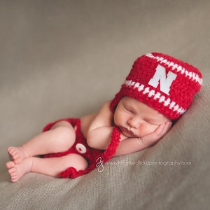 Puede incluir: Un bebé recién nacido durmiendo, con un gorro de ganchillo rojo y blanco con la letra "N" y un pañal a juego. El bebé está acostado sobre una manta de color neutro.