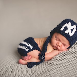 May include: A baby wearing a navy blue crocheted hat and shorts set. The hat has a white "NY" logo and ear flaps with tassels. The shorts have white stripes. The baby is lying on a grey blanket.