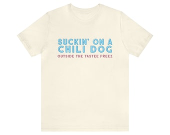 Könnte beinhalten: Ein cremefarbenes T-Shirt mit dem Text "Suckin' on a Chili Dog" in Blau mit rosa Umrandung. Der Text "Outside the Tastee Freez" steht in Rosa unter dem Haupttext.