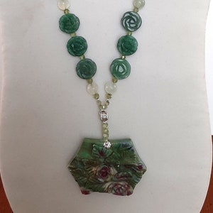 May include: A jade necklace featuring carved green rose-shaped beads and smaller round beads. The necklace has a pendant with a floral design and a silver-colored clasp. The necklace is displayed on a white mannequin.