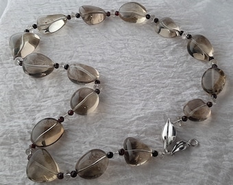 May include: A necklace featuring large, oval-shaped smoky quartz beads, interspersed with smaller red and clear beads. The necklace has a silver-colored leaf-shaped clasp. The beads have a translucent, light brown color.