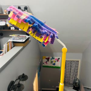 May include: A yellow duster with a multi-colored, fuzzy, fabric head. The duster is made of soft, fluffy material and is designed for cleaning.