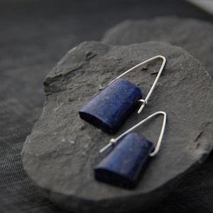 May include: A pair of silver earrings with dark blue rectangular lapis lazuli stones. The earrings are hanging from a dark gray stone surface.