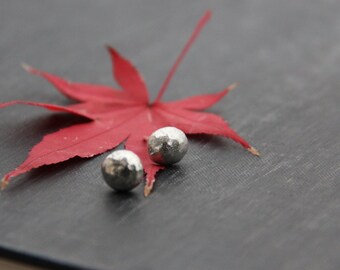 May include: Two silver hammered metal ball earrings on a grey surface with a red maple leaf.