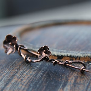 May include: A copper wire brooch with two flower-shaped accents. The brooch is on a wooden surface.