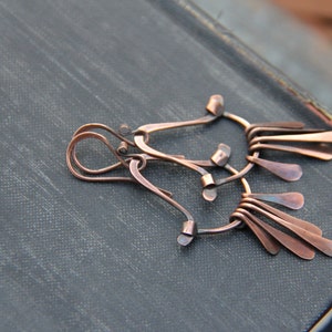 May include: A handcrafted copper-coloured metal earring with an abstract design. The earring features looped and curved elements, along with multiple teardrop-shaped dangles. The jewellery is set against a dark, textured background.