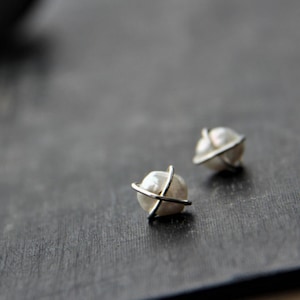 May include: Two small, pearl-like stud earrings with a silver-colored metal band encircling each. The earrings are set against a dark gray, textured background. The design is simple and elegant.