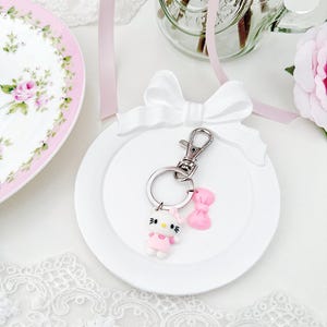 May include: A white ceramic dish with a white bow on top. The dish has a Hello Kitty keychain with a pink bow on it.
