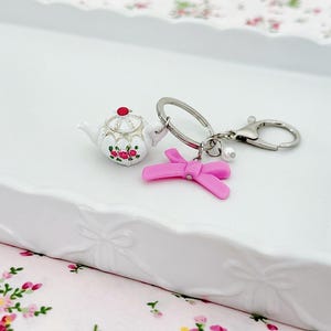 Pode incluir: Um porta-chaves prateado com um charm de bule branco com detalhes florais vermelhos e rosa, e um charm de laço rosa. O bule tem uma tampa vermelha e detalhes dourados. Sobre uma bandeja branca.