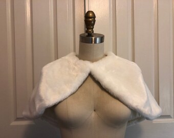 May include: White faux fur shrug with a shawl collar. The shrug is displayed on a white dress form.