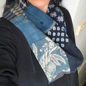 Può includere: Una sciarpa in patchwork blu e bianca con un motivo floreale. La sciarpa è realizzata con diversi tessuti ed è legata intorno al collo di una persona che indossa una camicia nera.