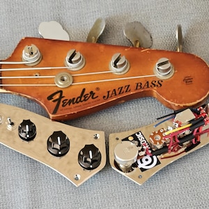 Op de afbeelding: Een vintage Fender Jazz Bass kop en bedieningspaneel. De kop is bruin met zilveren stemknoppen. Het bedieningspaneel is zilverkleurig met zwarte knoppen en elektronische componenten. De tekst "Fender Jazz Bass" is zichtbaar.