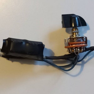 May include: A close-up of an electronic component, featuring a black knob atop a multi-position switch. The switch has a metallic finish with orange and silver accents. Wires are connected to the switch and encased in black insulation, with braided silver wires extending from the component.