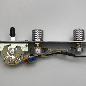 Puede incluir: Una placa de control de guitarra cromada con dos perillas texturizadas, un interruptor de palanca negro y un interruptor de múltiples posiciones. La placa tiene cableado y componentes electrónicos adjuntos. Hecho en los Estados Unidos.