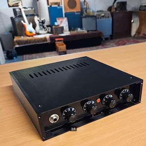 Op de afbeelding: Een zwarte audioversterker met vier bedieningsknoppen en een enkele ingang. De bovenkant van de versterker heeft een reeks ventilatiesleuven. De versterker staat op een houten oppervlak.