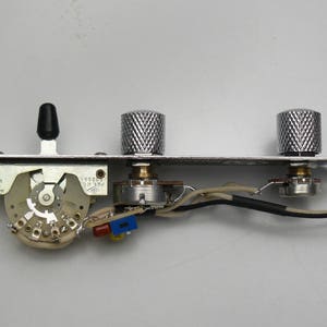 Peut inclure: Plaque de commande de guitare avec un interrupteur à bascule noir, deux boutons chromés et un commutateur multiposition. La plaque métallique comporte des composants électroniques et du câblage. Les boutons mesurent environ 2,5 cm de diamètre.