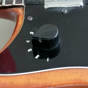 Puede incluir: Primer plano de la placa de control negra de una guitarra con una perilla negra y marcas numeradas blancas del 1 al 5. El cuerpo de madera de la guitarra es visible en el fondo.