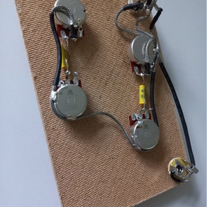 Può includere: Cablaggi per chitarra con quattro potenziometri, un interruttore e un jack, collegati da fili e condensatori. I componenti sono montati su una scheda rettangolare marrone chiaro strutturata. Il cablaggio è argentato e nero.